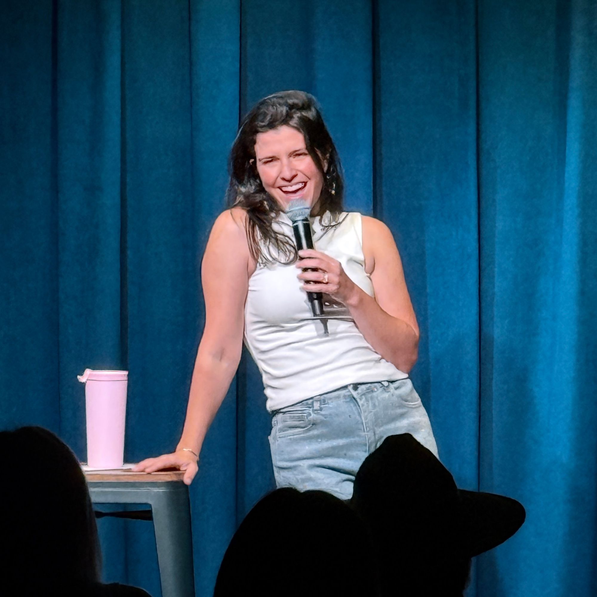 Comedian smiling on stage, leaning on a stool at the Snowmass Live Comedy Series, The Collective Snowmass