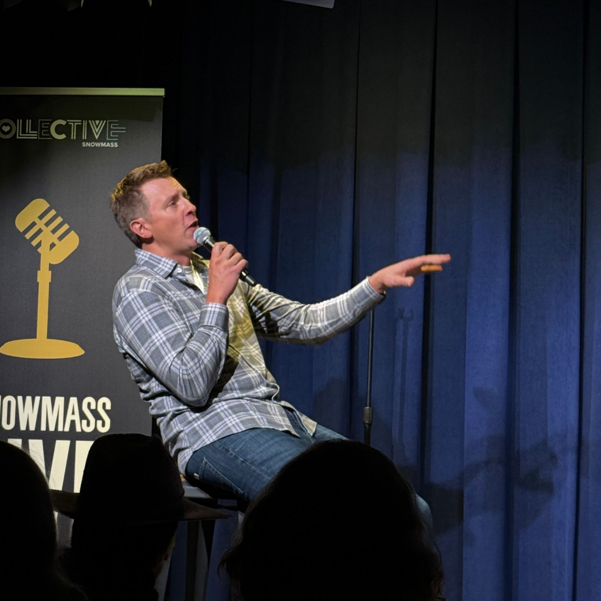 Comedian on a stool pointing to the crowd beside the Snowmass Live banner at the Snowmass Live Comedy Series, The Collective Snowmass