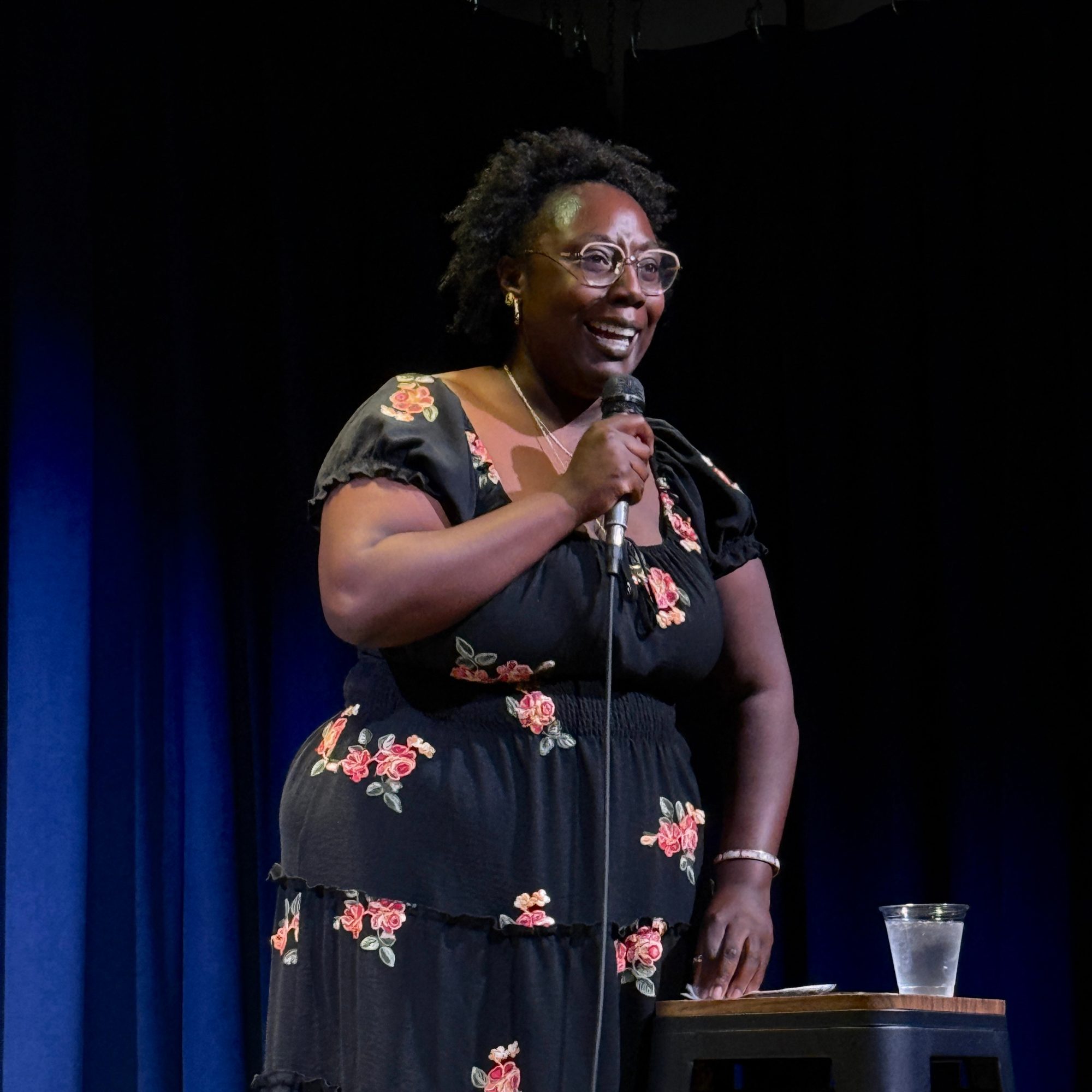 Comedian in a floral dress at the microphone stand at the Snowmass Live Comedy Series, The Collective Snowmass