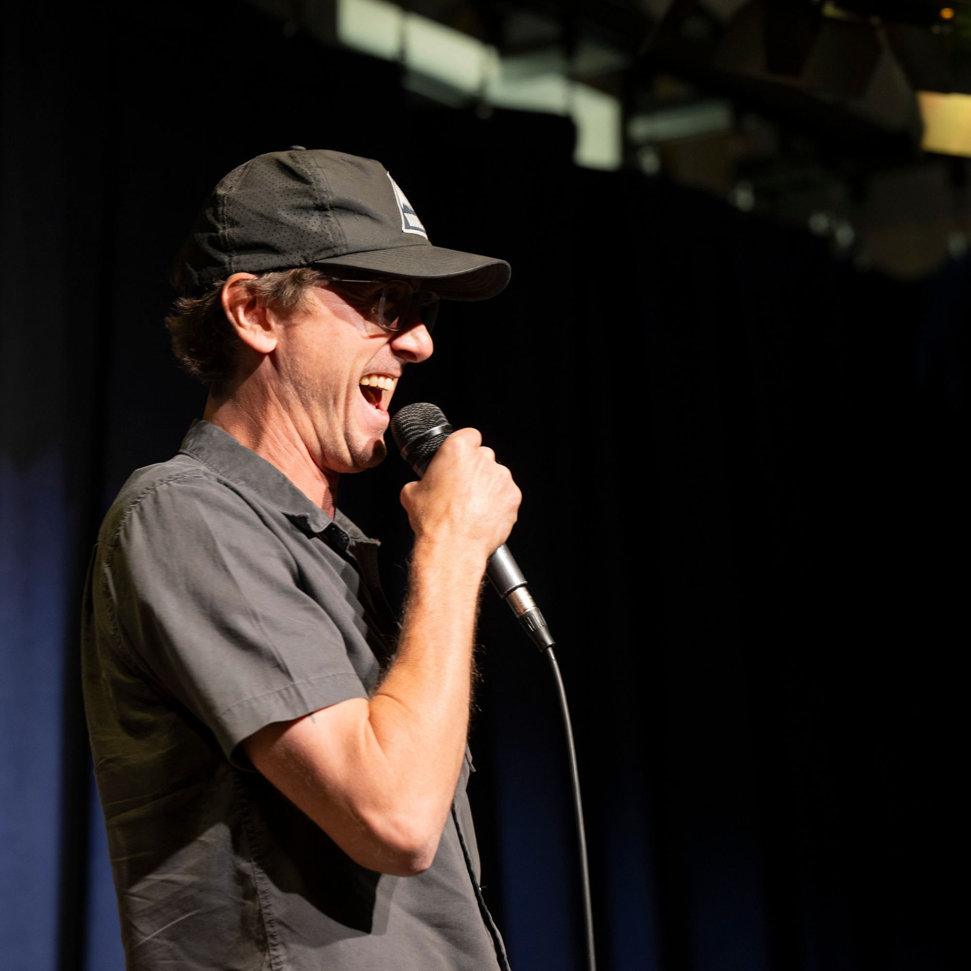 Comedian in a cap laughing into the microphone at Snowmass Live Comedy WKND!, The Collective Snowmass