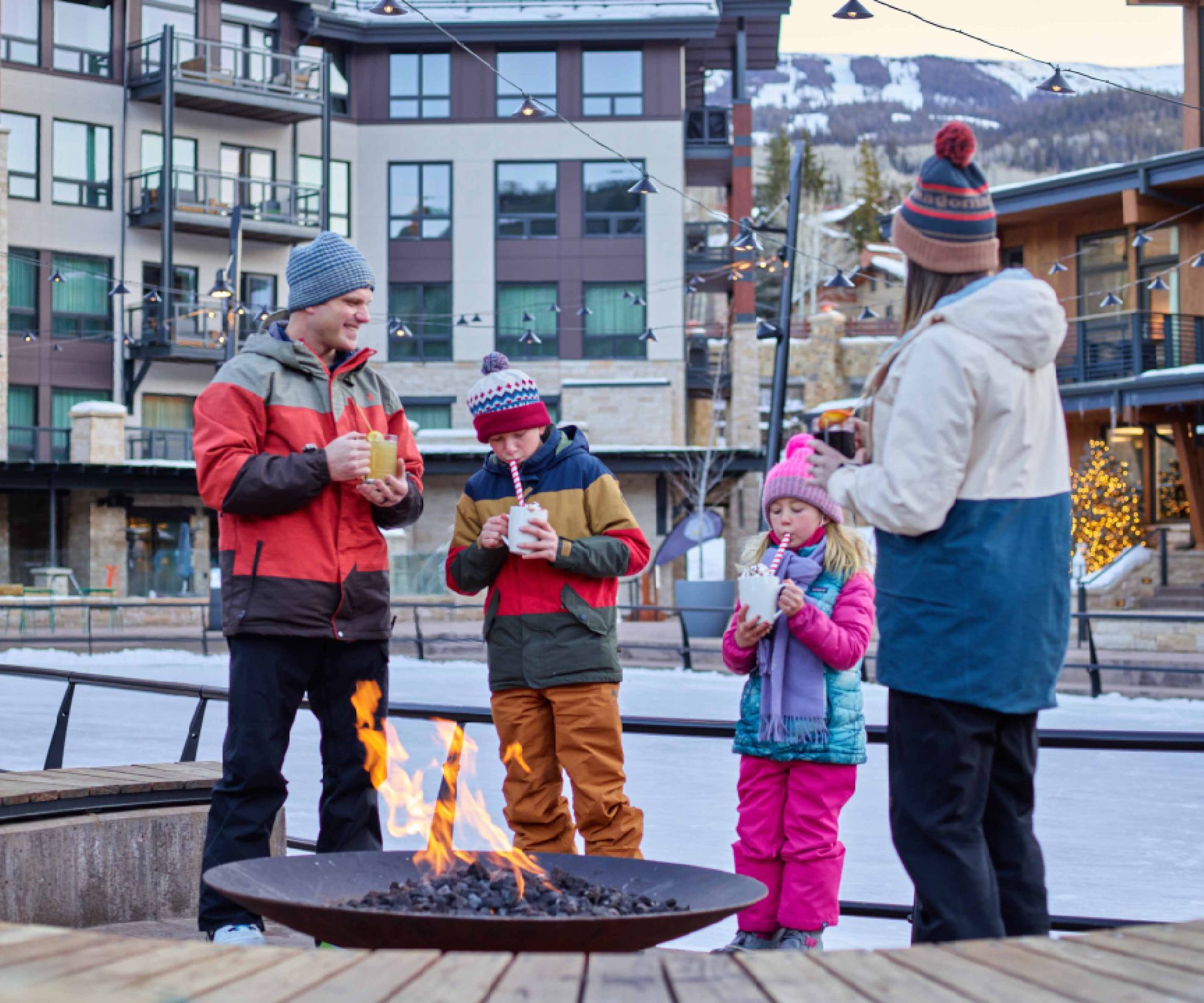 Family with hot drinks by the fire pit in Base Village, The Collective Snowmass